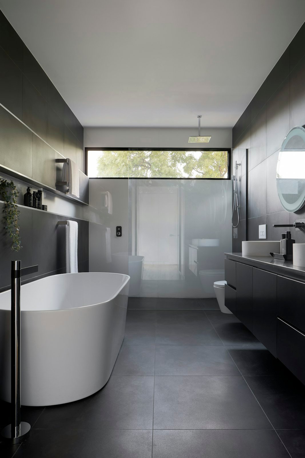 A dark stone bathroom with a freestanding tub under a high window.