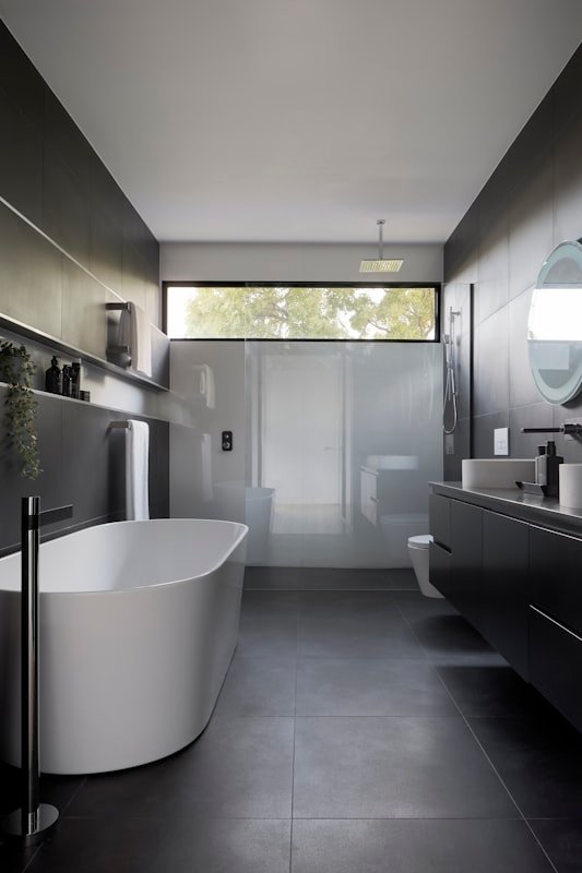 A dark stone bathroom with a freestanding tub under a high window.